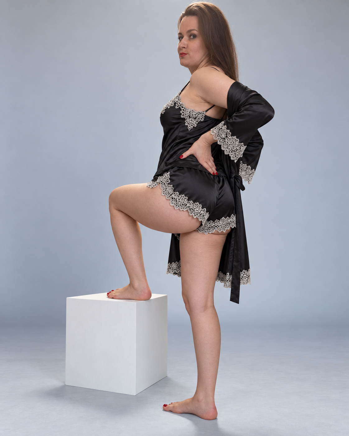 A woman models a black satin lingerie set with silver lace trim, consisting of a chemise and a matching robe, standing on a white block against a gray studio background.