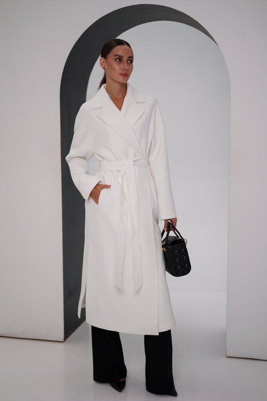 Woman wearing a white coat and black pants, holding a black handbag, against a plain background.