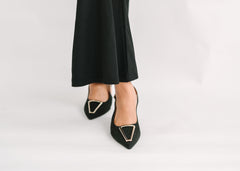 Black pointed-toe shoes with gold accents worn with black pants on a light gray background