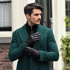 A man wearing a textured dark green double-breasted coat, black leather gloves, and a black zippered turtleneck, standing outdoors in front of a building with brick and white walls.