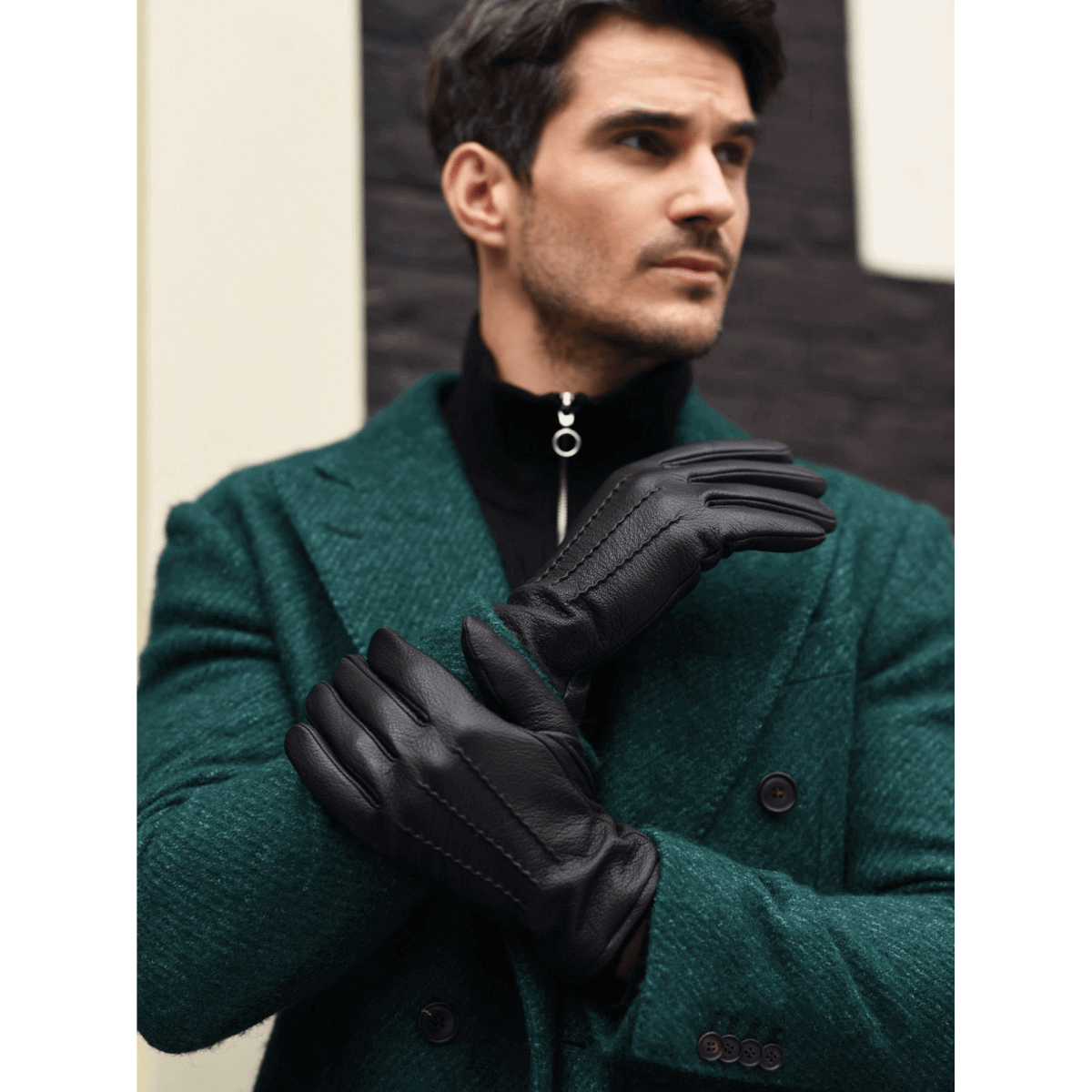 A man wearing black leather gloves with a quilted lining, styled over a dark green wool blazer and black zip-up sweater. He is posed against a modern architectural background with a dark brick pattern.