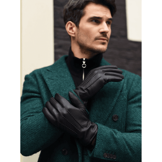 A man wearing black leather gloves with a quilted lining, styled over a dark green wool blazer and black zip-up sweater. He is posed against a modern architectural background with a dark brick pattern.