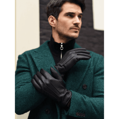 A man wearing black leather gloves with a quilted lining, styled over a dark green wool blazer and black zip-up sweater. He is posed against a modern architectural background with a dark brick pattern.