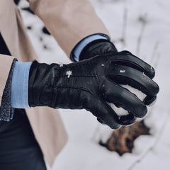 A close-up of a person wearing a black leather winter glove while holding snow in their other hand, with a beige coat and light blue shirt cuff visible.