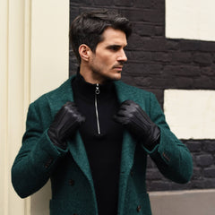 A man wearing a textured dark green wool-blend coat over a black zip-neck sweater, styled with black leather gloves, standing against a black and white brick wall.