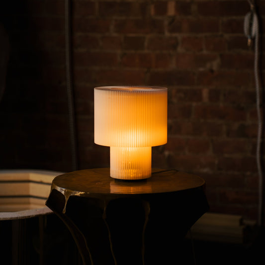Table lamp with ribbed design on a round wooden table against a brick wall.