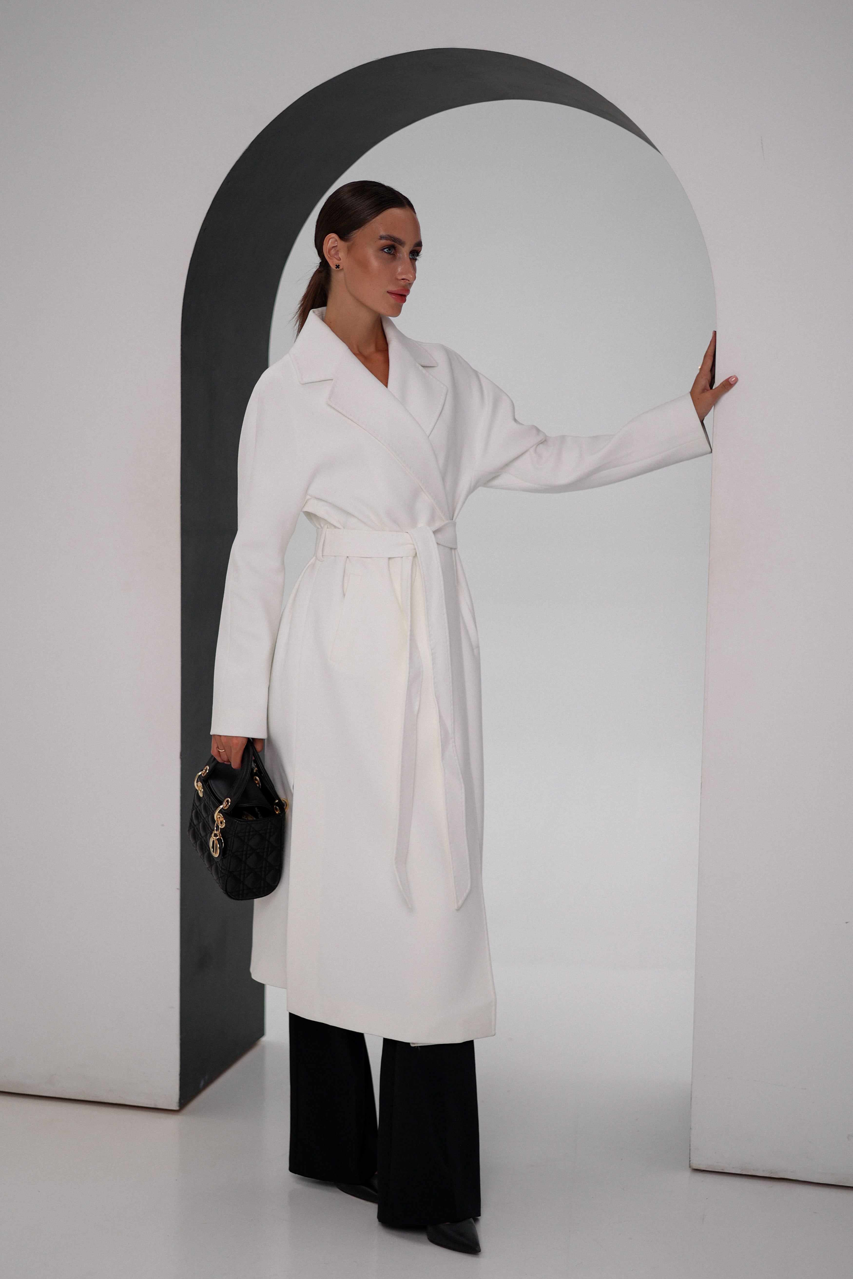 Woman wearing a white coat and black pants, holding a black handbag, against a plain background.