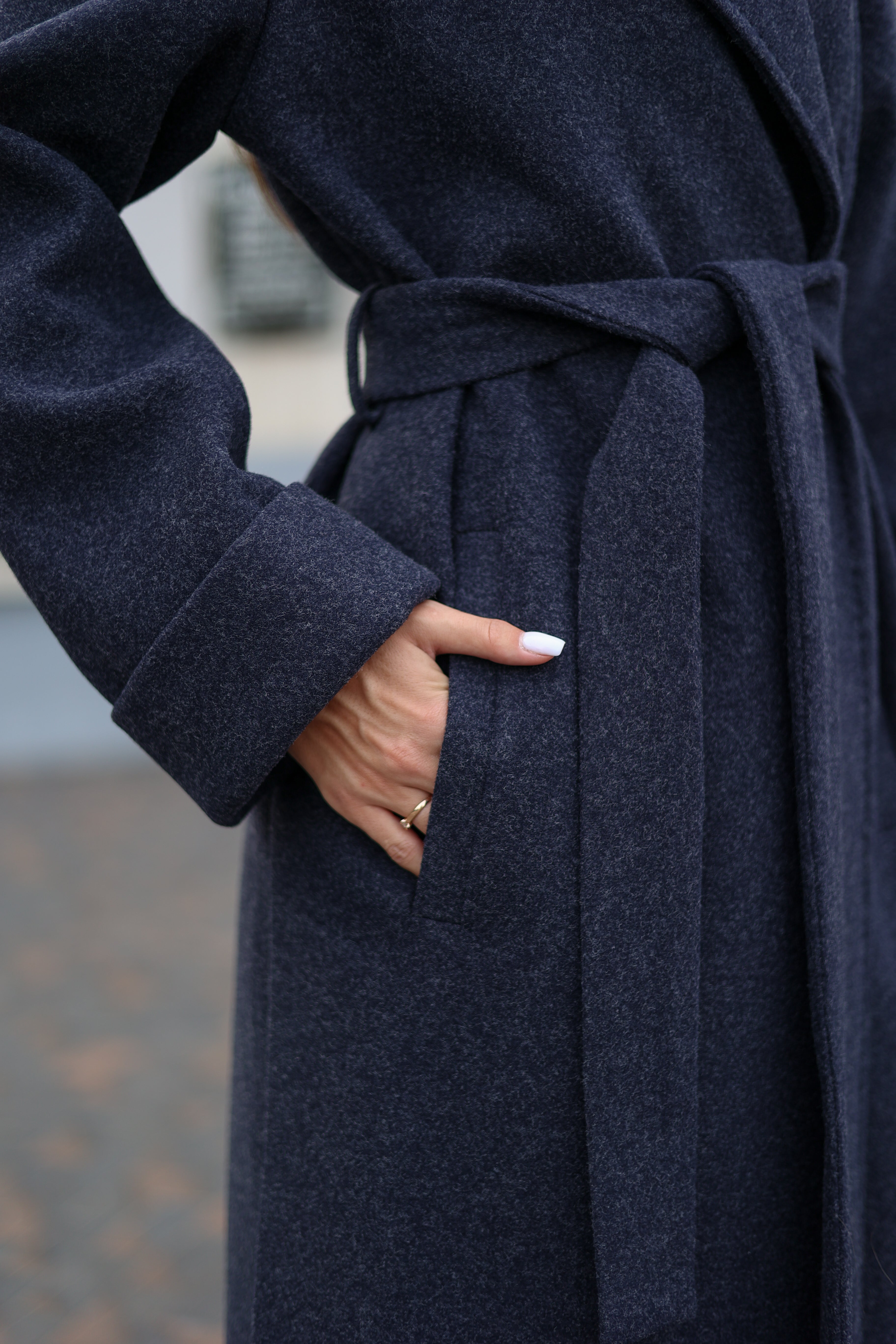 Woman wearing a long navy coat with a belt, holding a black handbag, standing on a paved walkway.