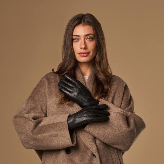 A woman wearing a brown wool coat and black leather gloves, posing with her arms crossed against a solid beige background.