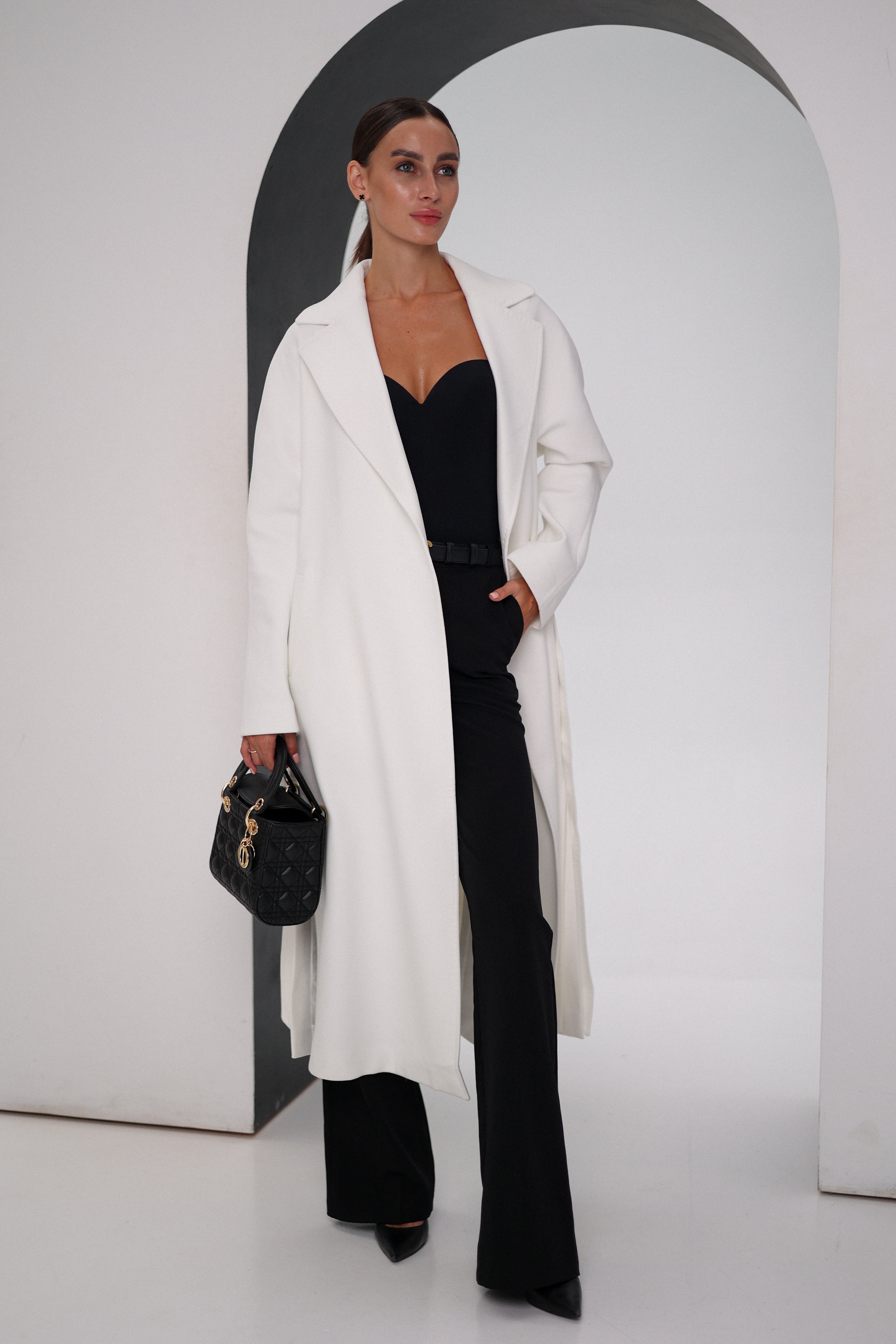 Woman wearing a white coat and black pants, holding a black handbag, against a plain background.