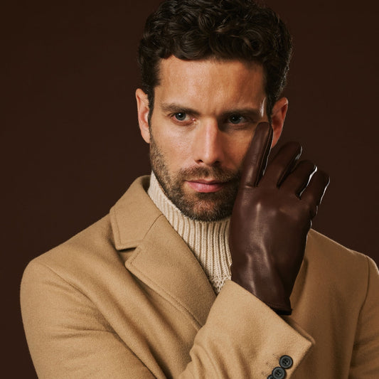 Man wearing a brown leather glove with a beige coat and white turtleneck against a brown background
