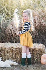 Young girl in a yellow dress and striped sweater standing next to pumpkins and hay bales.