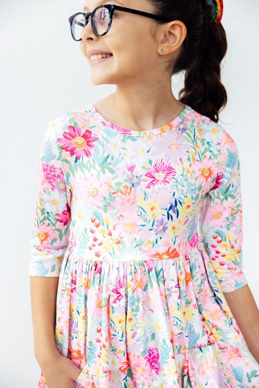 A young girl wearing a colorful floral print dress and black eyeglasses, smiling and looking to the side against a plain light background.