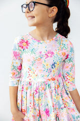 A young girl wearing a colorful floral print dress and black eyeglasses, smiling and looking to the side against a plain light background.