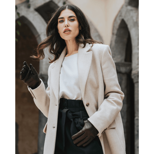 Woman wearing a beige coat and black gloves standing in front of a stone wall.
