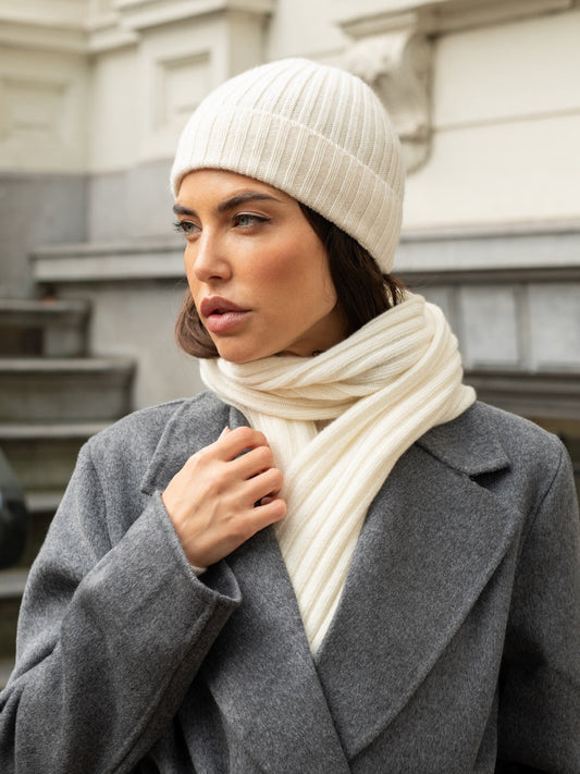 Woman wearing a white knit beanie and scarf with a gray coat outdoors.