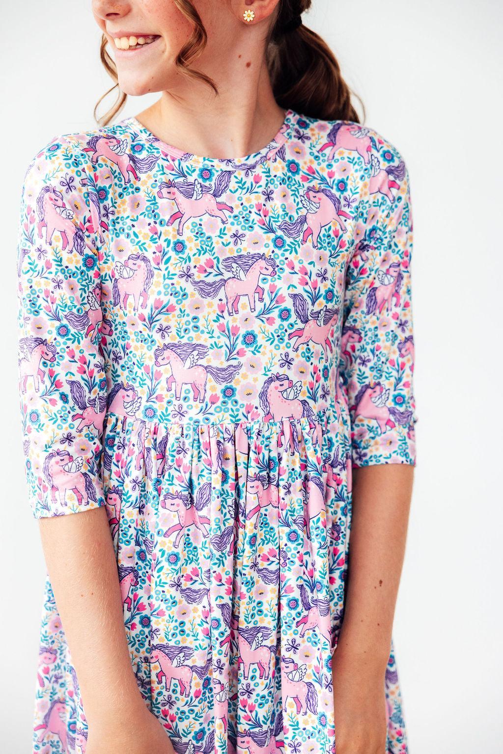 A woman wearing a pink and blue floral dress with a repeating unicorn pattern, shown from the chest down against a plain light background.