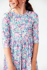 A woman wearing a pink and blue floral dress with a repeating unicorn pattern, shown from the chest down against a plain light background.