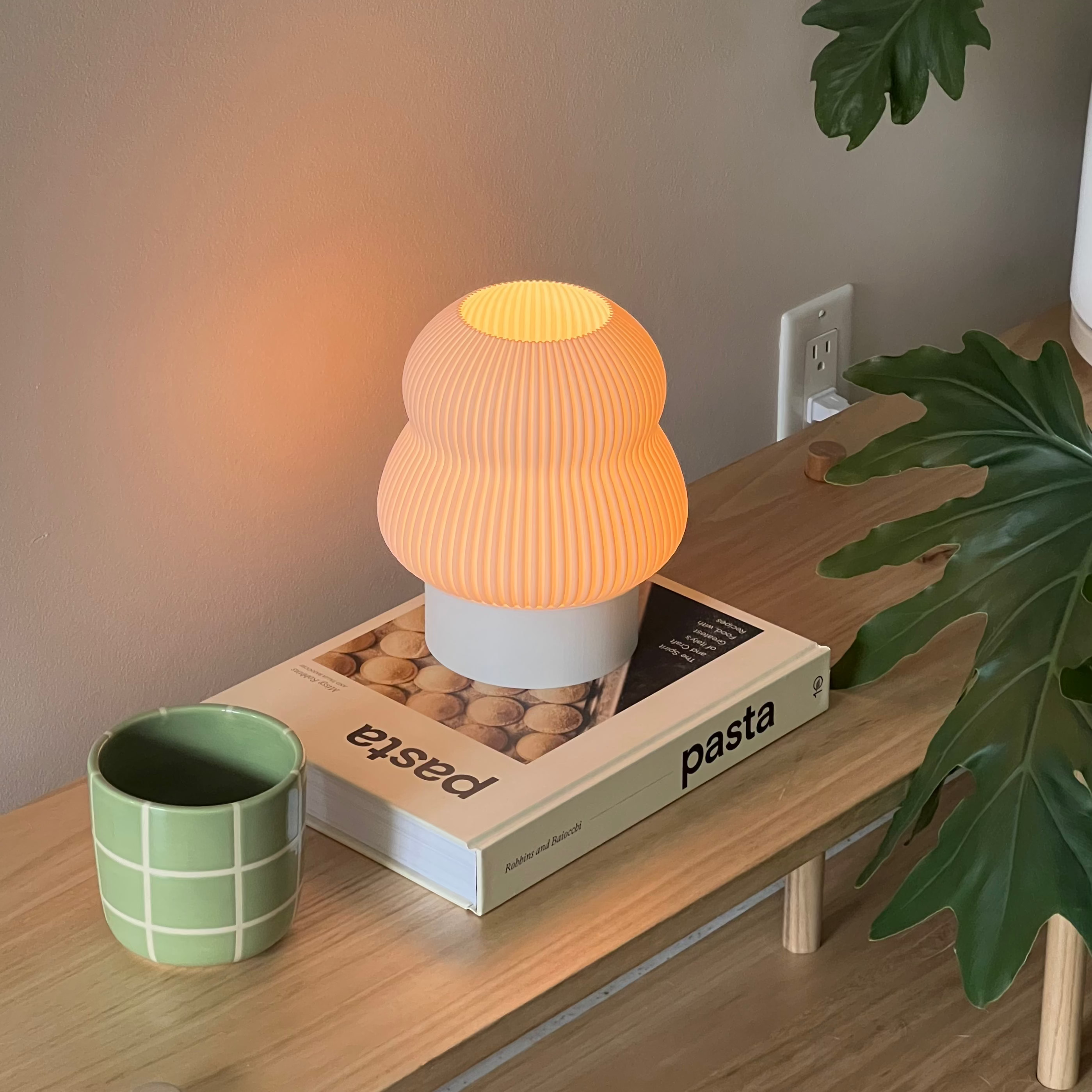 Small orange lamp on a box with 'pasta' text, next to a green mug and plant on a wooden surface.