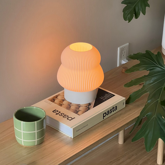 Small orange lamp on a box with 'pasta' text, next to a green mug and plant on a wooden surface.