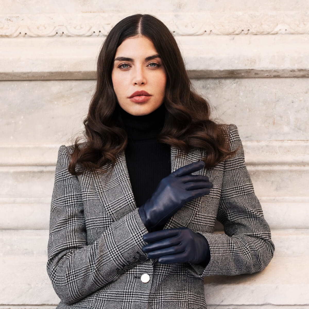 Woman wearing a gray checkered blazer and black gloves against a light stone wall.