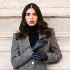 Woman wearing a gray checkered blazer and black gloves against a light stone wall.