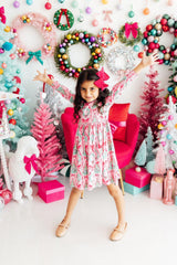 A young girl with a pink bow in her hair poses in a festive holiday dress with a pink and white pattern, surrounded by Christmas wreaths, decorated trees, and gift boxes.