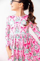 A young girl wearing a pink long-sleeve dress with a festive Christmas print of green trees, bows, and ornaments, shown from a side profile against a white background.