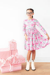 A young girl with glasses stands wearing a pink long-sleeve dress with a light blue and white pattern, paired with white cowboy boots. She is standing next to a stack of pink gift boxes on a light wood floor against a white wall.