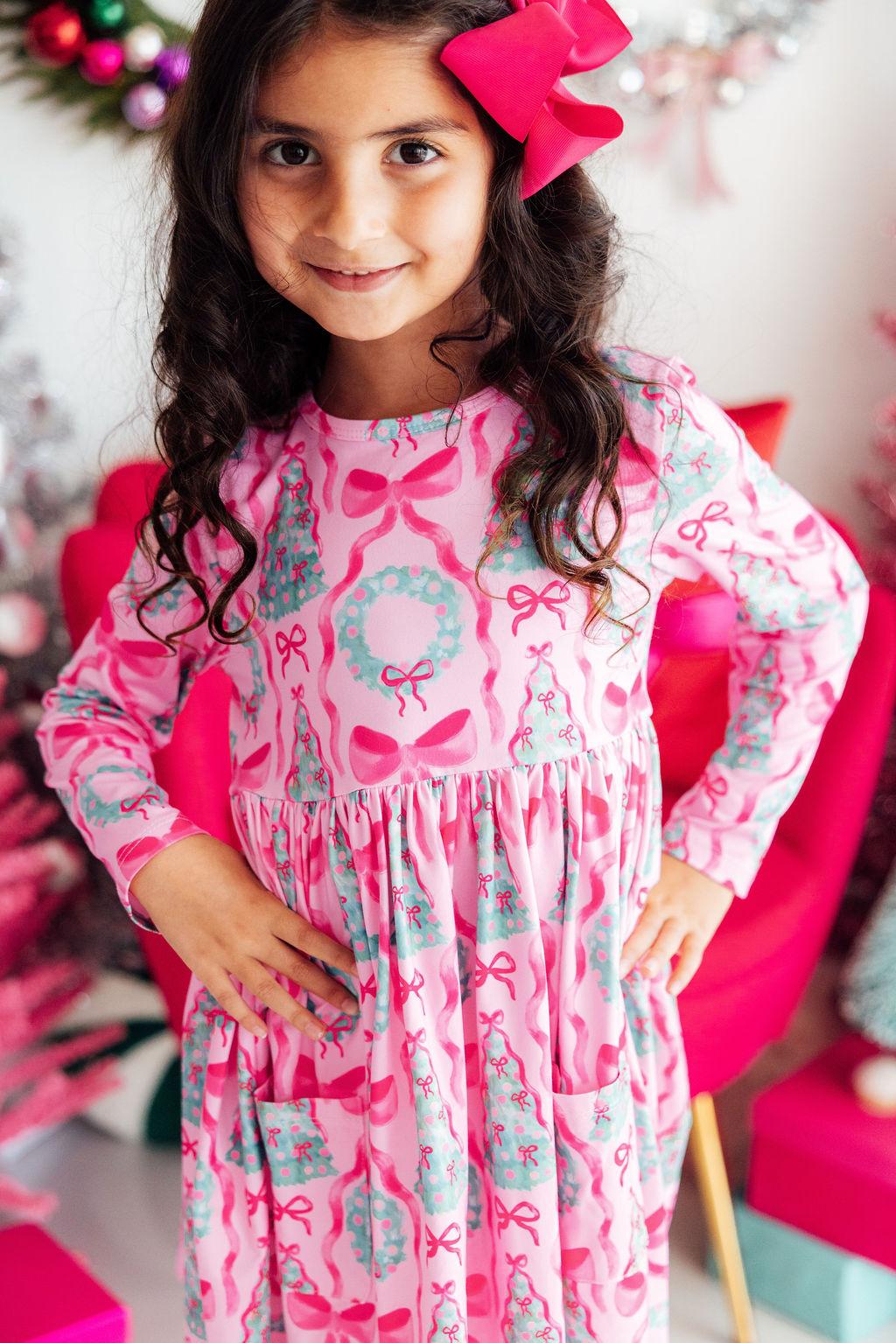 A young girl with long dark curly hair wearing a pink long-sleeve dress with a holiday print of bows, wreaths, and Christmas trees, accessorized with a large pink bow in her hair.