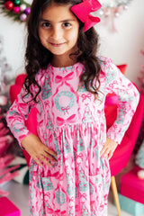 A young girl with long dark curly hair wearing a pink long-sleeve dress with a holiday print of bows, wreaths, and Christmas trees, accessorized with a large pink bow in her hair.