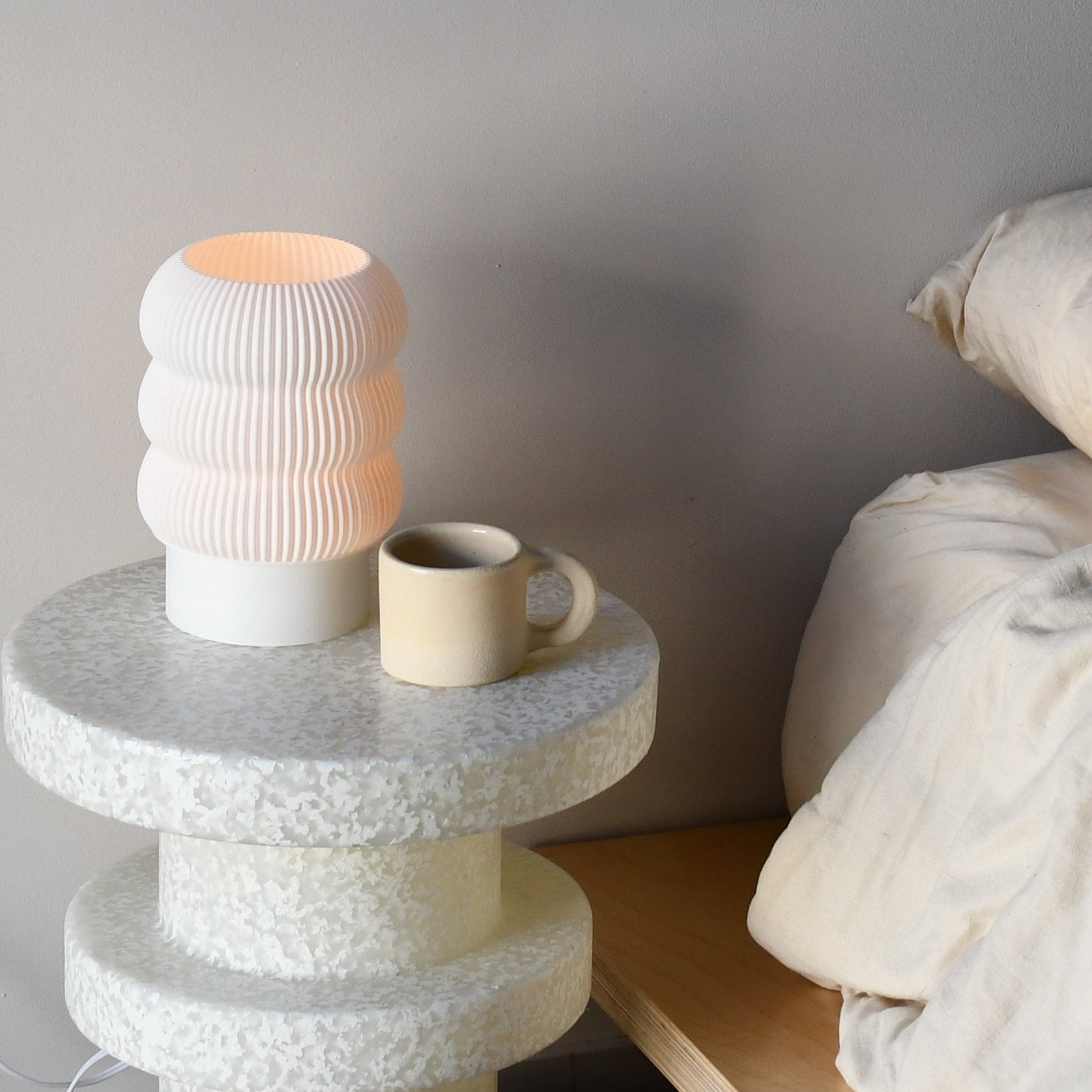 Small stone table with a textured lamp and mug next to a bed.