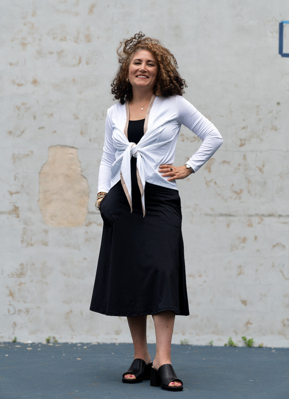 Woman wearing a white jacket over a black top and skirt against a plain wall.