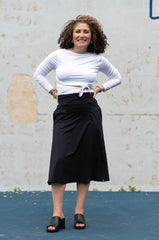 Woman wearing a white long-sleeve top and black skirt standing against a textured wall.
