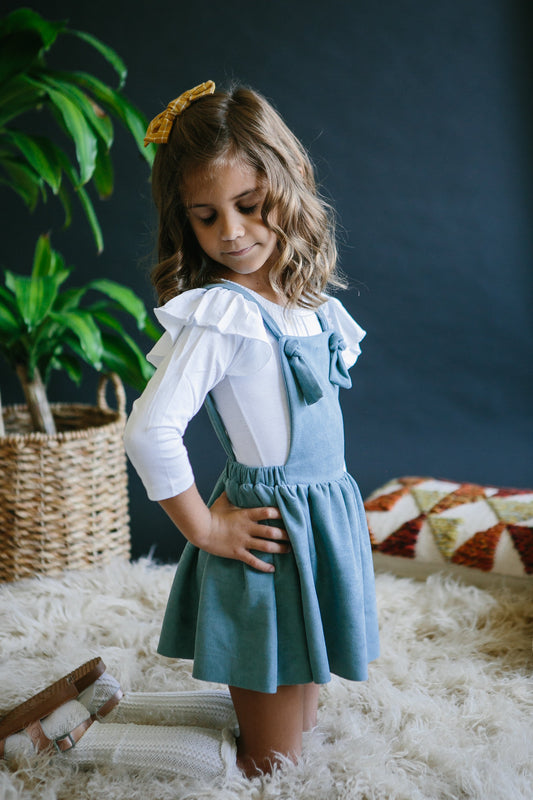 Young girl in a blue dress with a white shirt underneath, standing in a cozy room with plants and a rug.