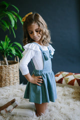 Young girl in a blue dress with a white shirt underneath, standing in a cozy room with plants and a rug.