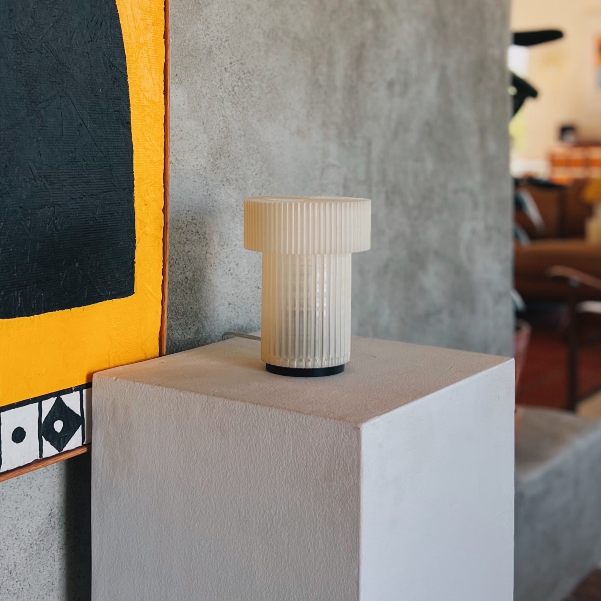 White ribbed vase on a white pedestal against a textured wall with abstract art.