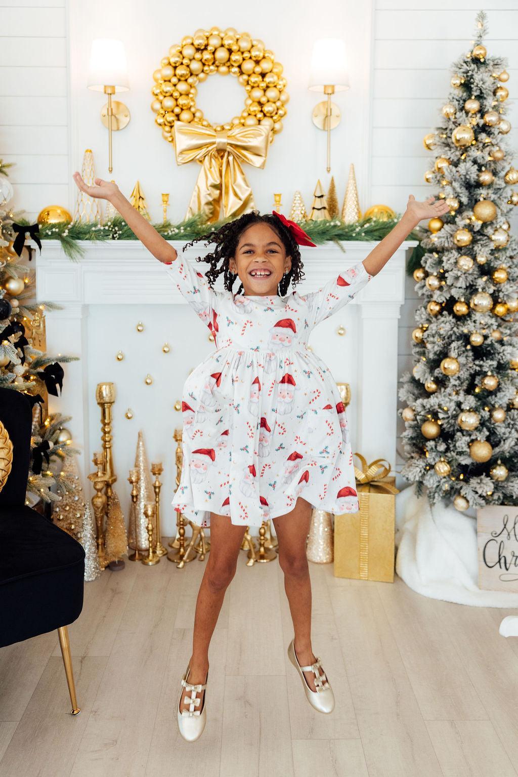 Child in a festive dress jumping in front of a decorated Christmas fireplace and tree.