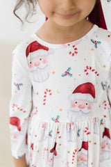 A close-up of a child wearing a white long-sleeve dress with a festive Christmas print featuring Santa Claus faces, candy canes, holly, and gold stars.