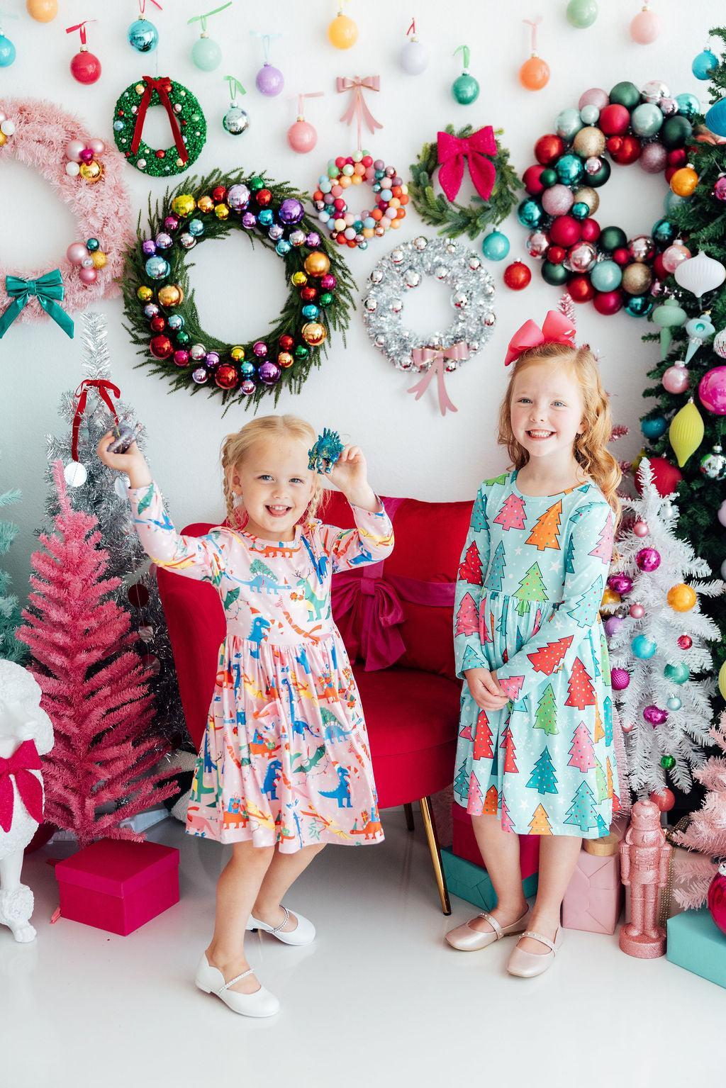 Two children in colorful dresses standing in front of a festive background with Christmas wreaths and decorations.