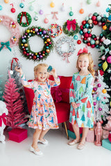 Two children in colorful dresses standing in front of a festive background with Christmas wreaths and decorations.