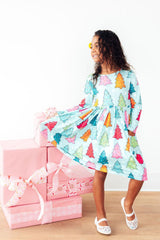 A young girl wearing a light blue long-sleeve dress with a colorful Christmas tree pattern, standing next to a stack of pink gift boxes on a light wood floor against a white wall.