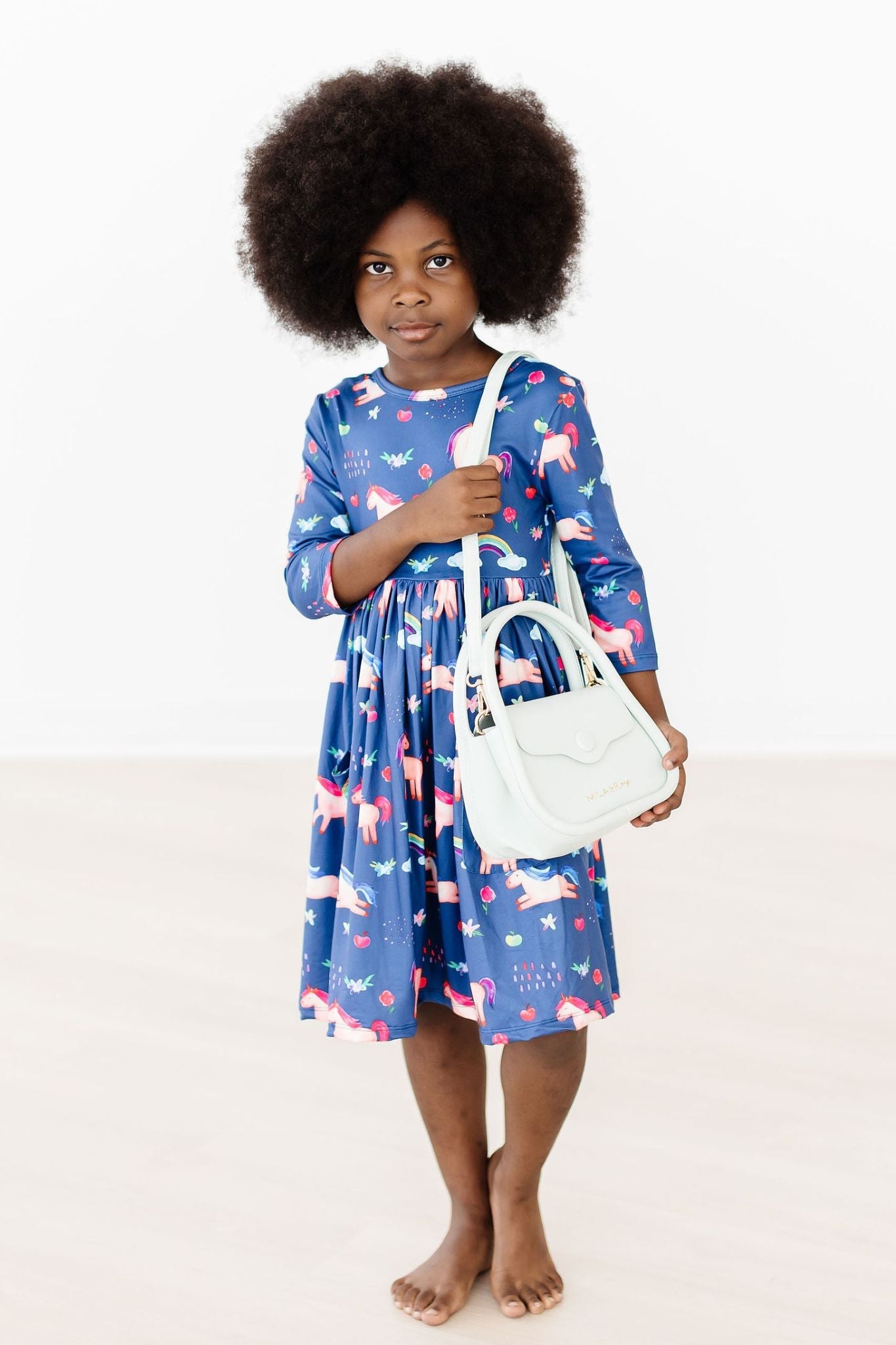 Child wearing a blue dress with pink floral pattern holding a white bag on a white background