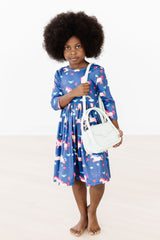 Child wearing a blue dress with pink floral pattern holding a white bag on a white background