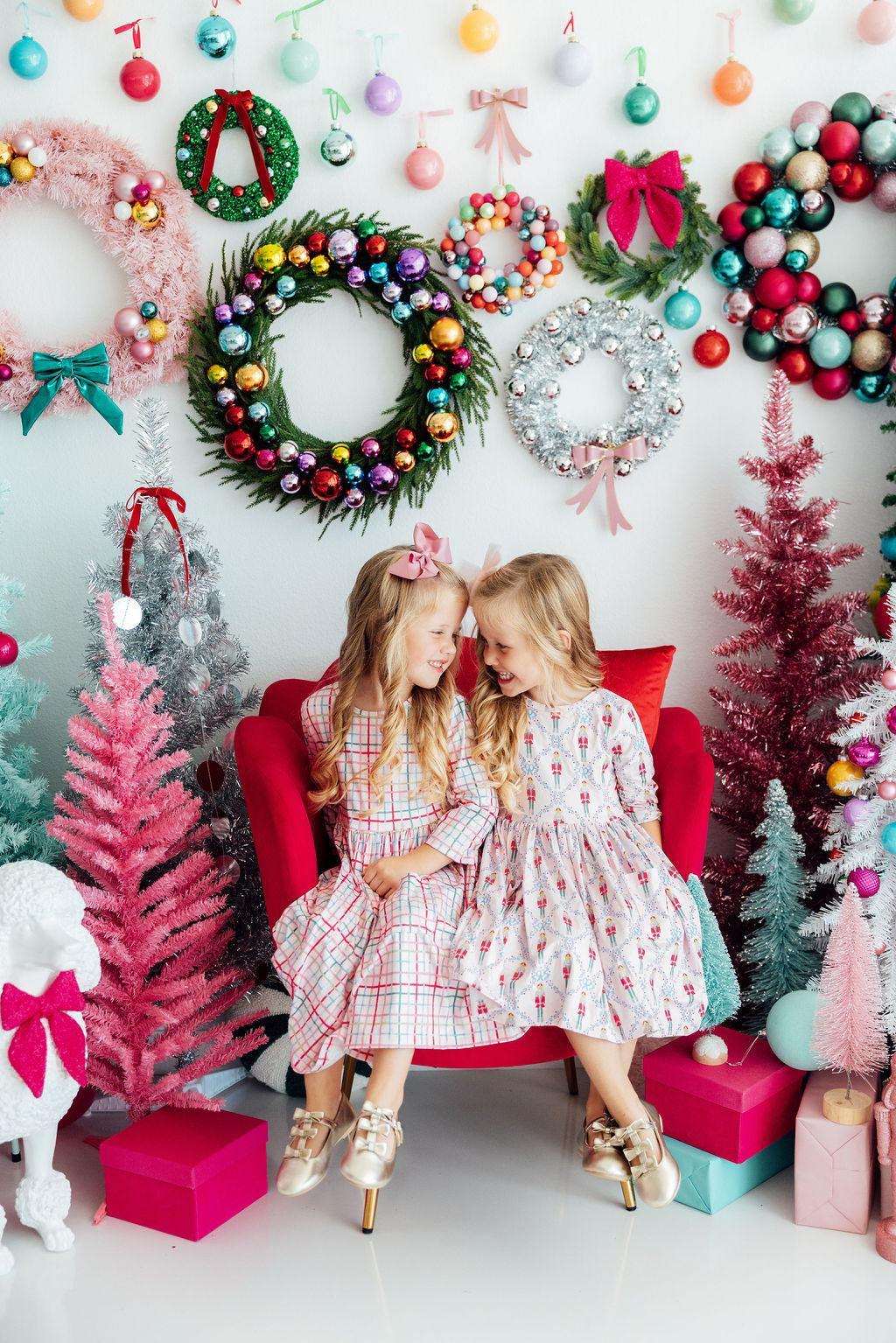 Two children sitting on a red chair surrounded by festive decorations including wreaths and Christmas trees.
