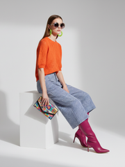 Woman in orange sweater, blue checkered pants, and pink boots sitting on a white cube with a colorful clutch.