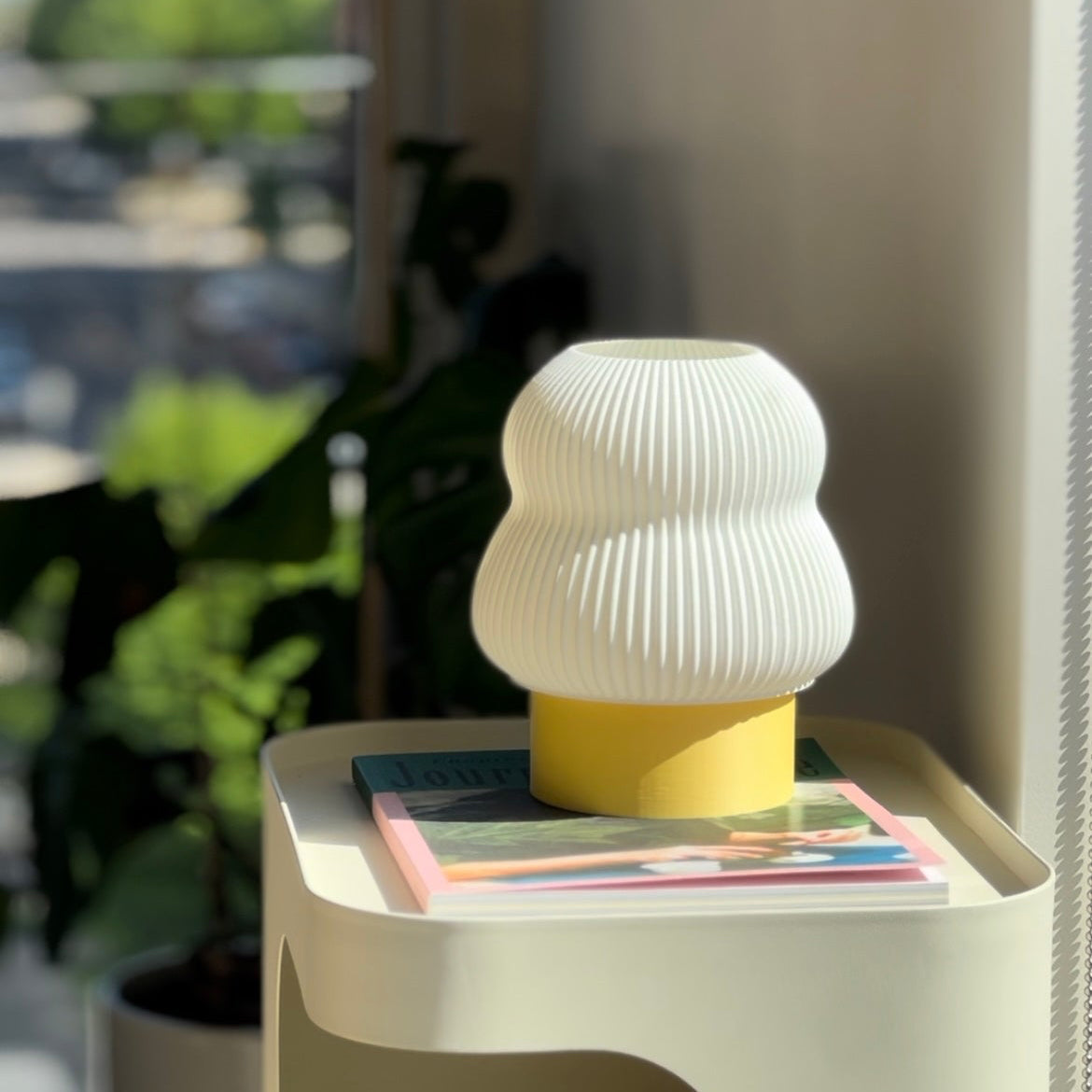 White textured lamp on a small table with magazines, blurred greenery in the background