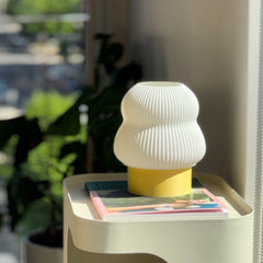 White textured lamp on a small table with magazines, blurred greenery in the background