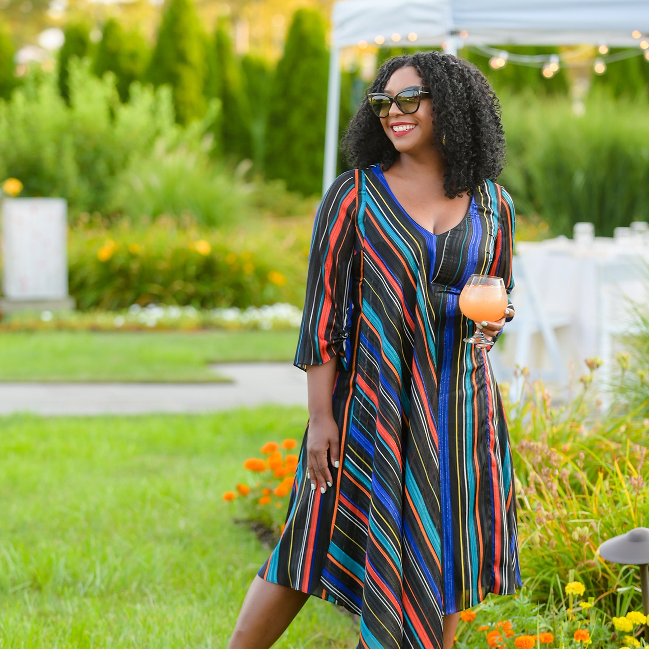 Woman in a colorful striped dress standing outdoors with greenery and flowers in the background