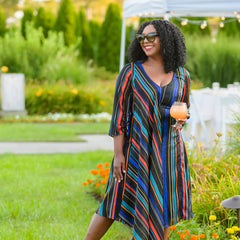 Woman in a colorful striped dress standing outdoors with greenery and flowers in the background